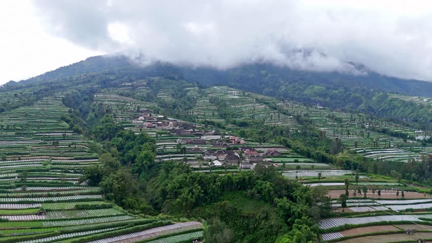 Indonesia rural landscape on mountainside. Aerial view of village on the mountain slope surrounded by plantation in foggy weather