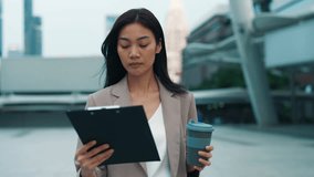 Portrait of serious Thai businesswoman with coffee cup checking report walking on clipboard outdoors. Asian brunette entrepreneur reads business papers on street. Paperwork control - Powered by Shutterstock - Get 15% off with code: PIKWIZARD15