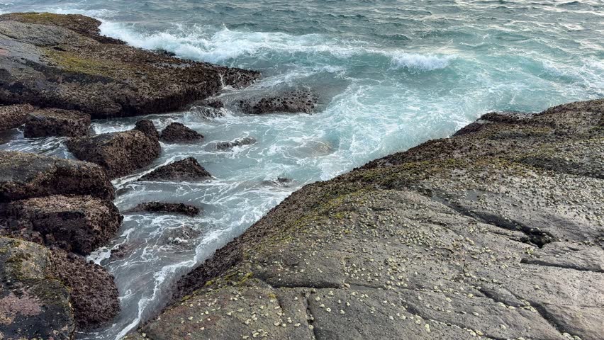 A rocky shoreline with a large body of water in the background. The waves are crashing against the rocks, creating a powerful and dynamic scene. The rocks are scattered throughout the area