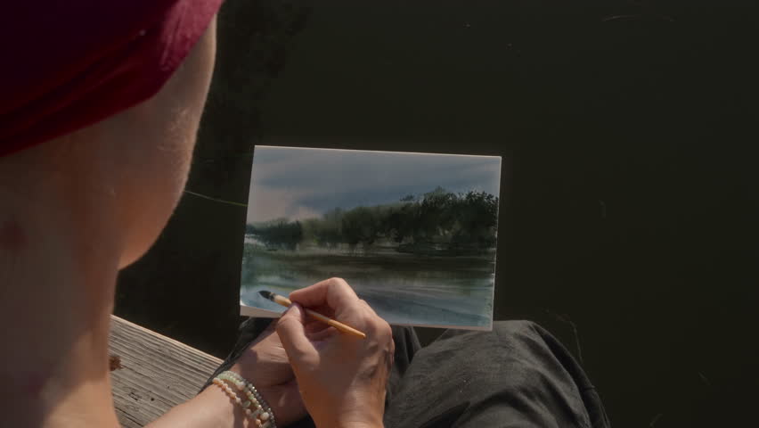 Female artist painting landscape sitting on wooden pier on river bank