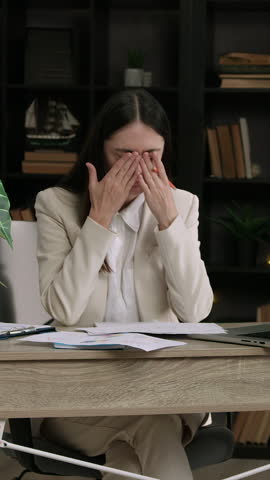 A lot of work for an important project. A brunette woman in a suit sits at an office table and slowly massages her temples and eyes, having a headache from fatigue and a lot of concentration at work.