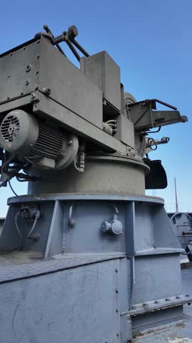 Close-up of a deck-mounted naval gun with targeting sights and ship radar mast in background, on a decommissioned gray military vessel under a blue sky.