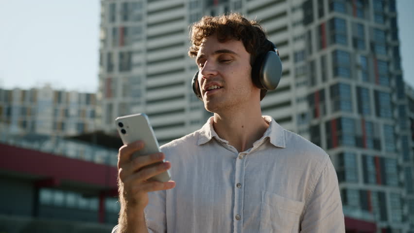 Happy positive Caucasian man holding mobile phone smartphone listening music in headphones guy enjoying outdoors relaxing weekend student carefree male businessman smiling city urban song app business