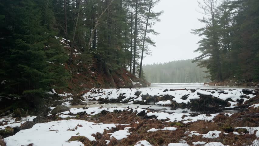 The snow is covering a beaver dam that separates a lake surrounded by forest in a misty winter day in Algonquin Park, Ontario, Canada