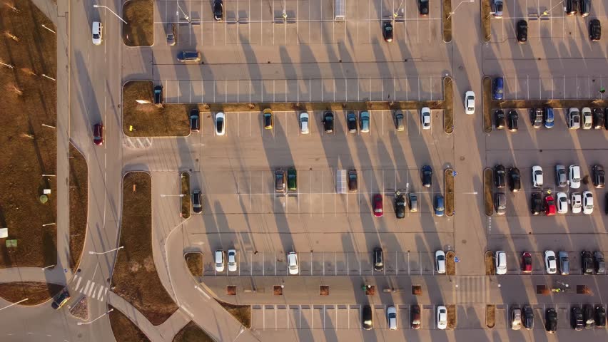 Aerial overview of a busy parking lot at sunset, long shadows stretching across parked cars and lanes, tracking backdrop