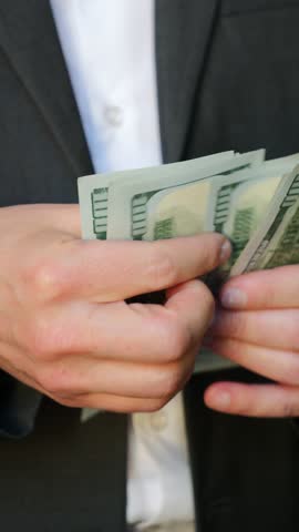 Businessman in a suit counts several hundred dollar bills in a bright outdoor setting.