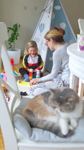 A mother and child playing with colorful building blocks in a cozy home, with a cat sleeping on a chair in the foreground. A warm and loving family moment filled with joy and creativity.