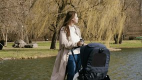 Young and happy mother walks in the park with the stroller and baby and drinks a coffee.Woman enjoys a coffee and time with her baby on maternity leave on a sunny windy day in the forest near the pond - Powered by Shutterstock - Get 15% off with code: PIKWIZARD15