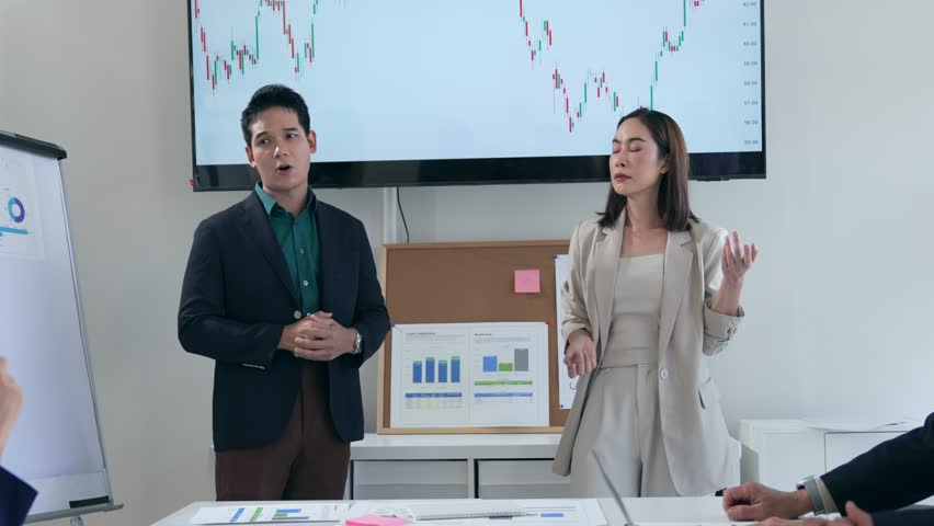 Two business partners, a man and a woman, present financial results to their team in a modern meeting room, using a large screen displaying stock market charts - Powered by Shutterstock - Get 15% off with code: PIKWIZARD15