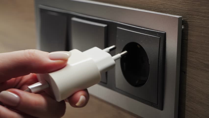 Close-up footage of hand inserting electrical plug into wall socket, connecting power source for electronic devices. 