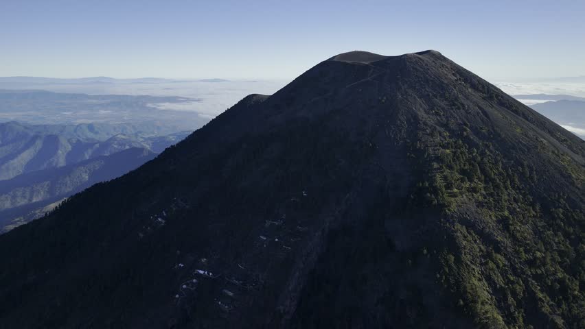 Drone orbits around dark side of Acatenango Volcano with a sunlit valley covered in clouds and the Agua Volcano on the skyline in the distance