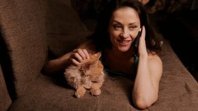 Smiling emotional woman speaking on mobile phone and stroking with love the hand her cute orange maine coon cat, lying on sofa at home interior. Closeup relax emotional indoor video. Vintage color - Powered by Shutterstock - Get 15% off with code: PIKWIZARD15