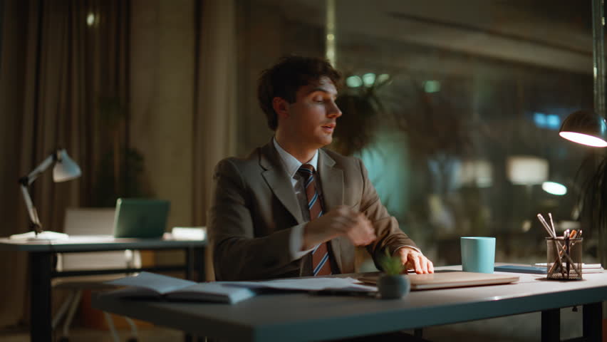 Tired man working overtime sitting dark workspace closeup. Exhauster sleepy businessman looking documents falling asleep in night office. Guy startuper close laptop lid drinking coffee having deadline