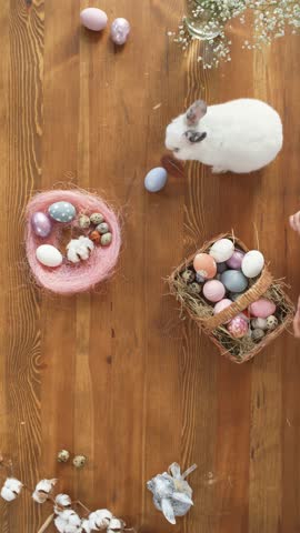 Overhead 4K vertical video clip of Easter egg nest on wooden table. A white rabbit grooms itself as a woman's hands organize decorated Easter eggs into a wicker basket. Easter holiday theme video.