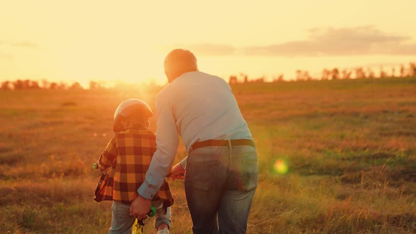 Father teaches child wearing safety helmet to ride bicycle. Family in park. Child rides bicycle. Father helps his son ride bike. Boy Kid, dad play together, sunset. Child dream learns to ride bicycle