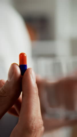Female fingers delicately grip a bright orange and blue capsule pill, preparing to take medication