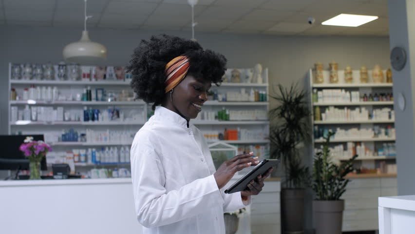 Female pharmacist using digital tablet while working at pharmacy counter, managing healthcare technology with confident smile