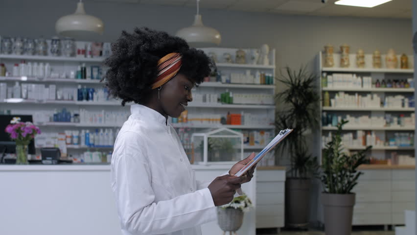 Professional african american female pharmacist navigating digital tablet while working at modern pharmacy counter, providing patient care with friendly demeanor