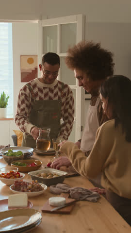 Vertical shot of three friends cooking festive meal together, chopping vegetables and pouring drinks for friendly home gathering
