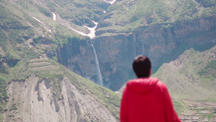 Defocused Indian male tourist standing in front of Sissu waterfall during the summer season and looking towards it at Sissu in Lahaul Valley, Himachal Pradesh, India. Tourist looks at scenic view.
