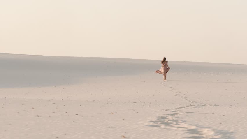 A graceful woman in a flowing pink dress walks through expansive white sand dunes, creating footprints in the serene landscape. The cinematic scene conveys beauty, solitude, and timeless elegance.