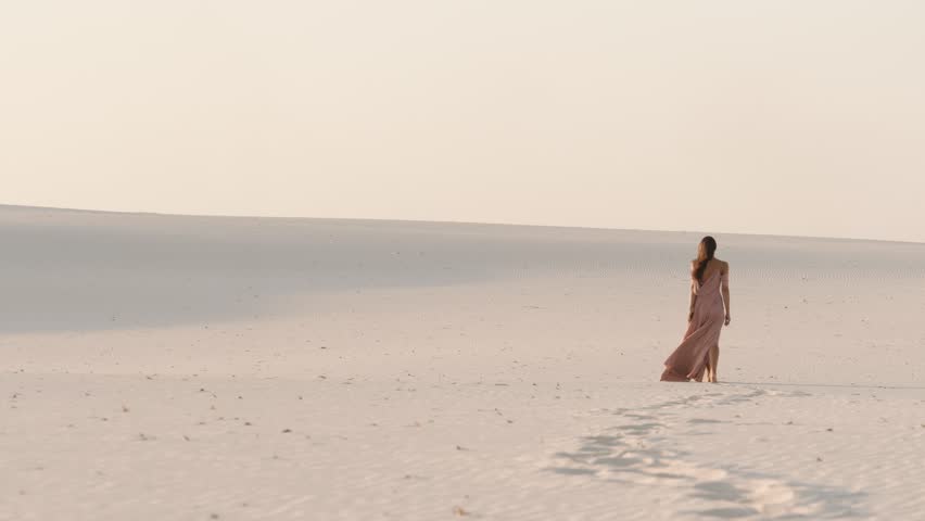 A stunning woman walks gracefully across expansive white sand dunes, leaving a trail of footprints. The epic scene features soft lighting, vast landscapes, and slow-motion elegance, ideal for
