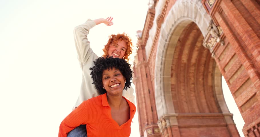 Two happy multi-ethnic female friends piggybacking in front of the arc de triomf in barcelona, spain