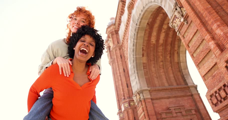 Two friends are enjoying a piggyback ride and laughing under arc de triomf in barcelona