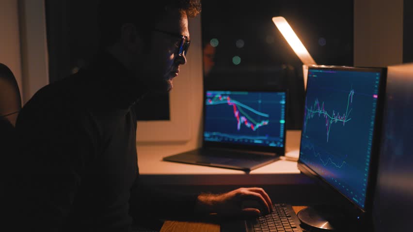 In a dimly lit room, a man focuses on his work, analyzing stock market data on two computer screens. He appears dedicated, wearing glasses and concentrating on trading activities.