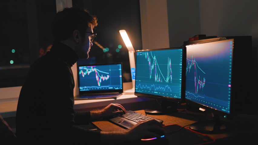A trading expert studies stock market trends in a cozy home office at night. Multiple monitors display live graph data while he focuses intently on his analysis.
