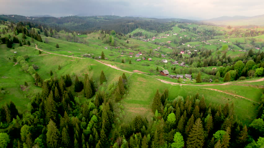 Aerial photograph of serene rural landscape in Ukraine, featuring rolling green hills, winding dirt roads, and scattered houses. Dense forests surround area, with dramatic, cloudy sky overhead.