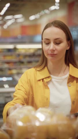 In a bustling store, a woman inspects a tray of freshly made donuts. She carefully scans the treats, contemplating her choices while surrounded by vibrant displays and shoppers.