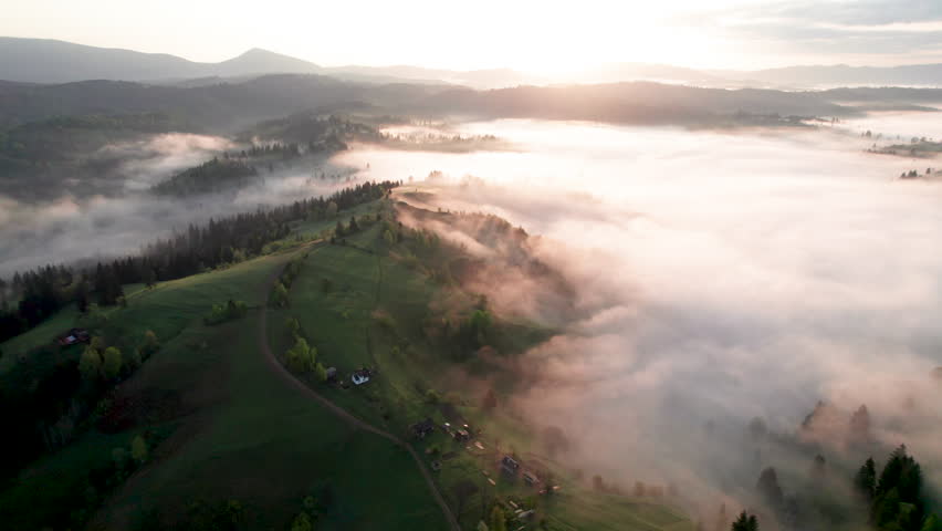 Aerial view of sunrise over mist-covered landscape, with golden light illuminating rolling hills and valleys. Thick fog creates mystical atmosphere, partially obscuring lush greenery and trees below.