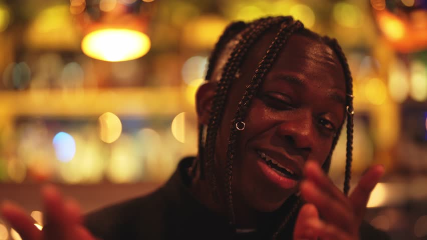 Portrait of charismatic young African American man talking cheerfully in bar. Handsome black male rapper flowing his rap, talented and perspective rap performer, stylish male hairstyle with braids
