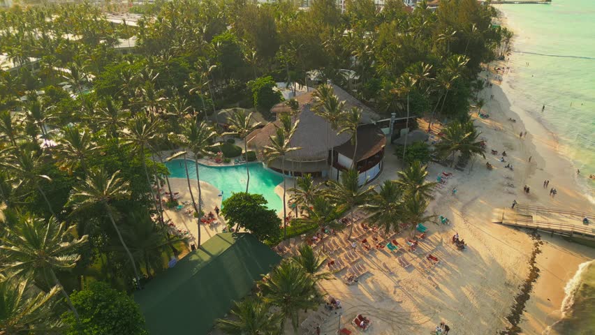 Aerial view of a tropical beach at the luxurious all-inclusive Impressive hotel in Punta Cana. The sunset casts a warm, bright light over everything. Tourists are relaxing and swimming in the pool