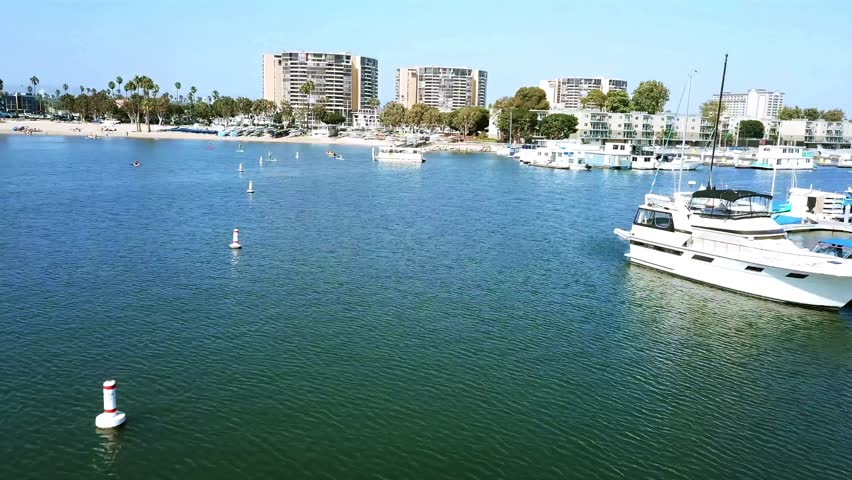 A view from drone flying over the ocean with boats, approaching Mother's Beach with greenery and urban skyline on a sunny day in Long Beach, CA.