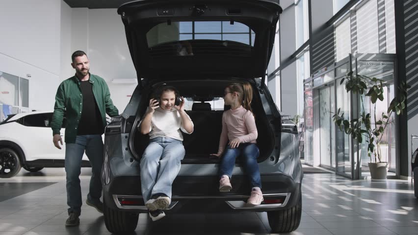 A happy father with his two daughters sits in the trunk of the car he just bought. The happy family is enjoying the purchase. The end of a test drive or purchase of a new, modern car.