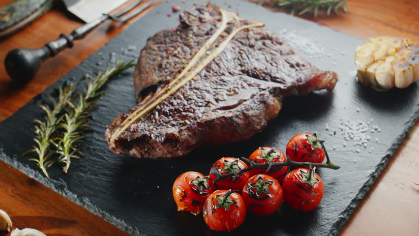 T-bone steak with herbs and garlic and tomatoes on the serving slate board close-up. Top view.
