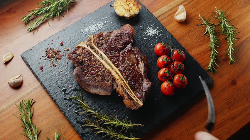 Sliced medium-rare T-bone steak with herbs and garlic on the serving slate board close-up.