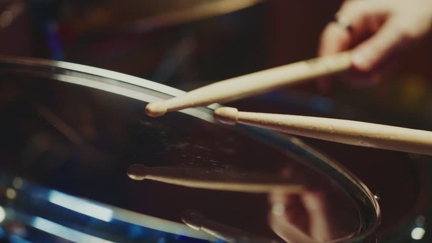 A close-up of a drum stick poised above a snare drum, highlighting the anticipation of a live music performance. The atmosphere is electric with energy from the crowd.
