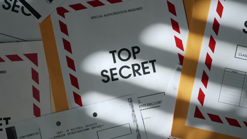 Close up on Top Secret label on manila envelope with classified documents. Dark office room and desk with window shadow on stack of papers with sensitive information and censored text