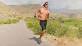 Shirtless athlete in cap, sunglasses, and headphones runs down the road at a steady pace, moving to his music as summer sunlight illuminates the path through green plains and mountain slopes. - Powered by Shutterstock - Get 15% off with code: PIKWIZARD15