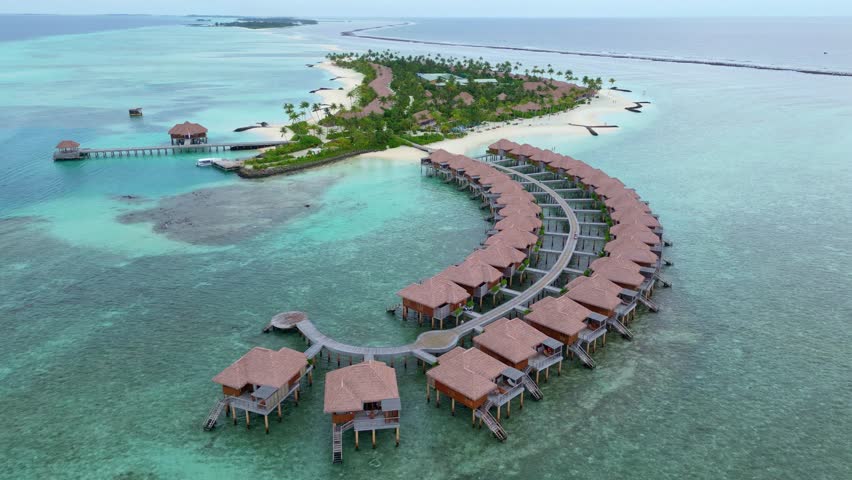 Exclusive and noble hotel villas surrounded by blue clear ocean water. Maldives Island with Luxury Water Villa Bungalows. Aerial Backwards wide shot.