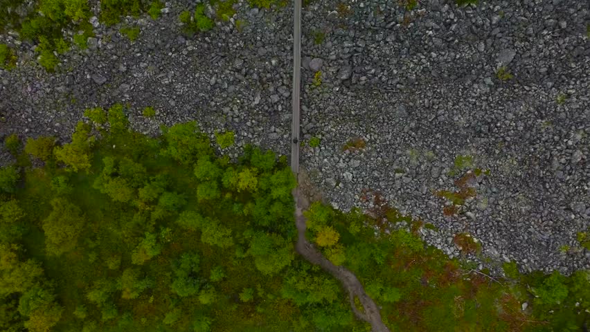 Top down aerial drone footage of a person walking on a wooden hiking trail or a bridge over some rocky and granite gravel covered terrain during cloudy day at Sweden. green forest trees on the bottom.