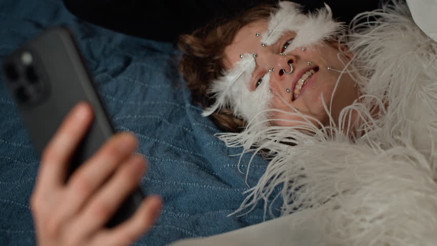 Tilt up shot of two cheerful female friends dressed in theatrical outfits with creative makeup filming smartphone video when lying on bed during home costume party