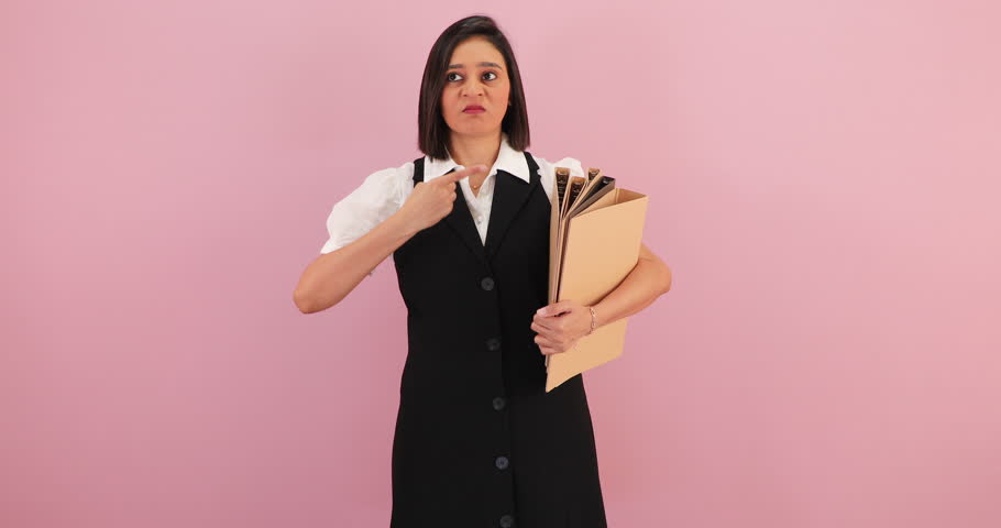 Unhappy and angry young South Asian woman pointing with furious to the folders she's holding and trying to stop her anger, isolated on pink backdrop
