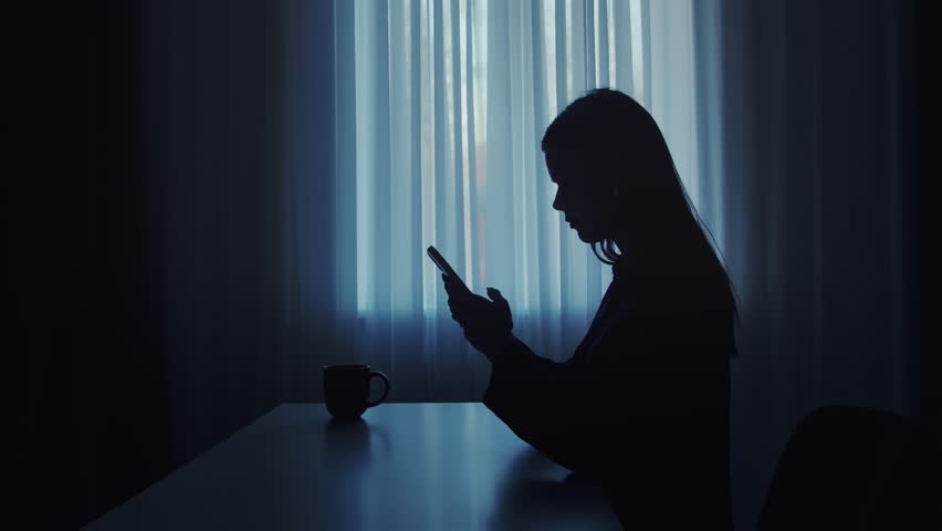Silhouette of a woman sitting at a table in front of a window, using a smartphone with a cup nearby.