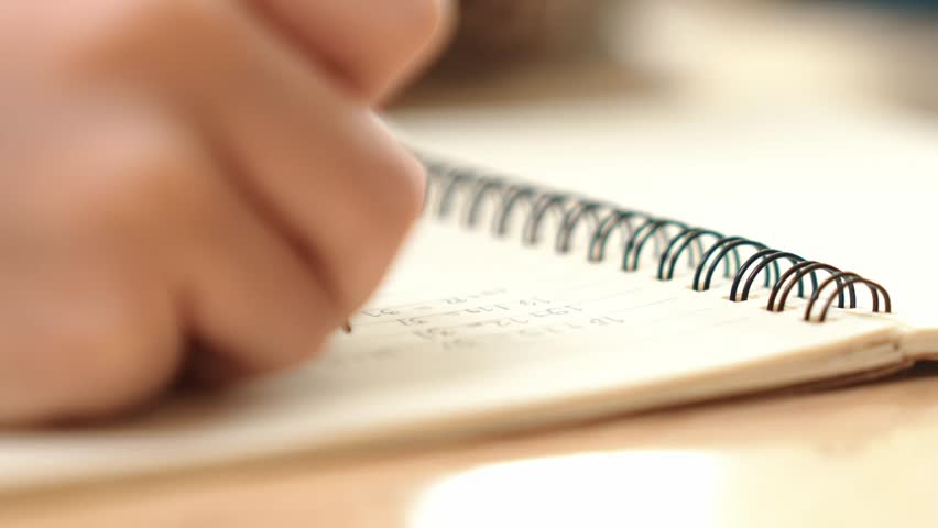 Close up of child hand taking a note or writing a letter at classroom. Happy student hands hold pen at book or doing homework or doing test at elementary school. Focus on hand. Education. Pedagogy.