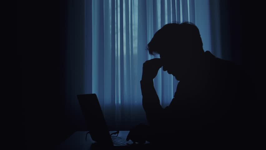 A silhouette of an individual typing on a laptop in a dimly lit room, creating a mysterious atmosphere.