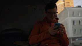 Young male passenger wearing orange jacket and glasses, smiling while using smartphone during bus journey, looking out window and enjoying urban commute - Powered by Shutterstock - Get 15% off with code: PIKWIZARD15